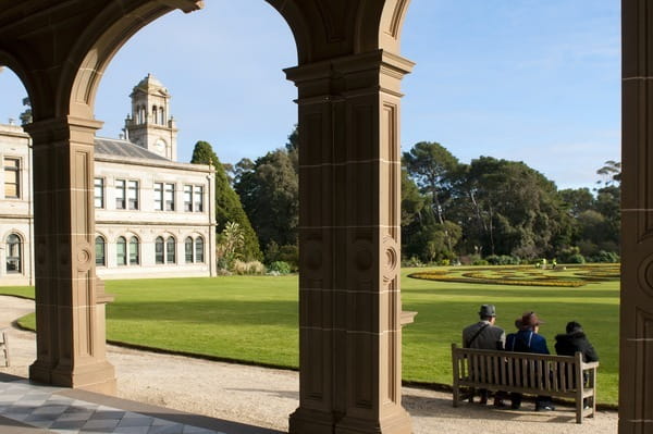 Three people sitting on a bench with Werribee Mansion in the background.