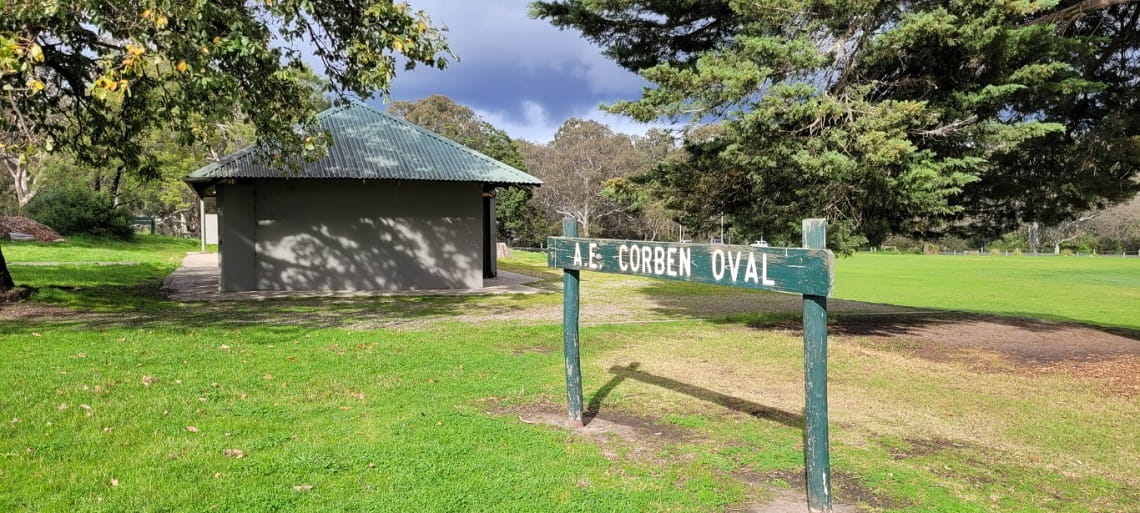 A grey and green building is in the distance under the shade of trees. A sign that reads 'Corben Oval' stands in the foreground.
