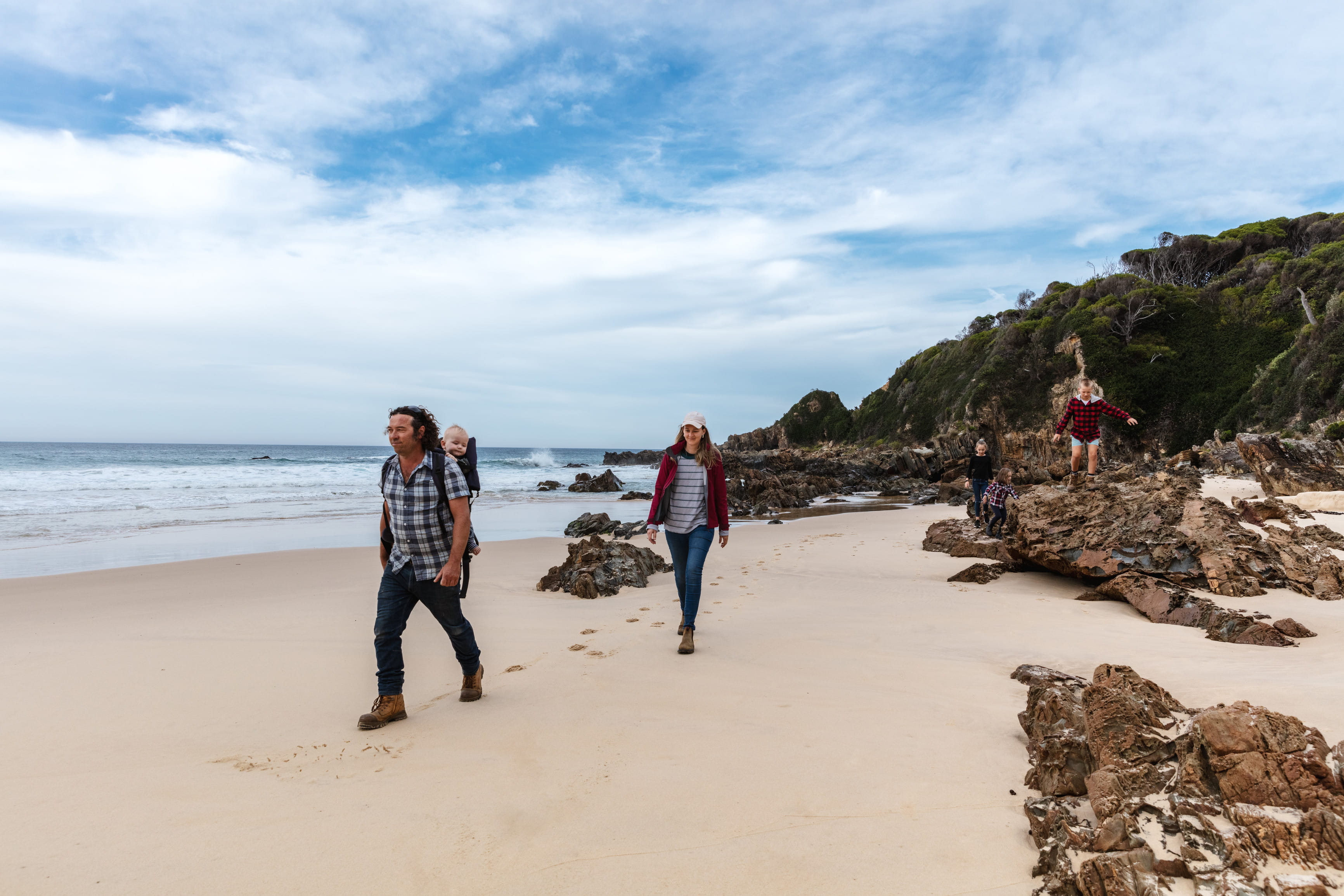 A man, woman, two children and a baby in a backpack walk along the sand on a rock strewn beach