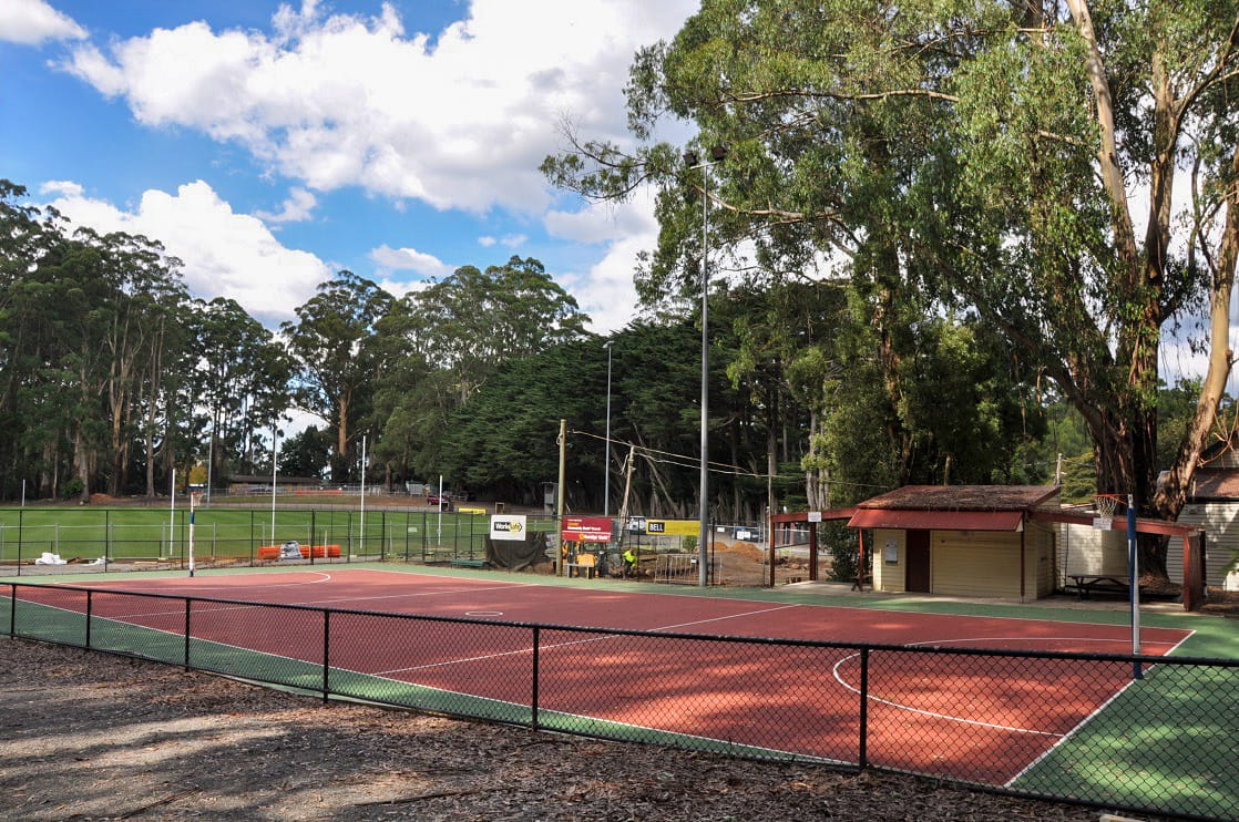 The existing netball court at the Olinda Recreation Reserve. Its surface is red and green in colour, and it is surrounded by a construction fencing and tall trees. It is adjacent to the oval.