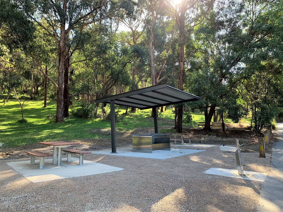 A new picnic area, with picnic tables on the left, an open shelter in the middle with a stainless steel barbecue underneath it, and a stainless steel drinking tap to the right.