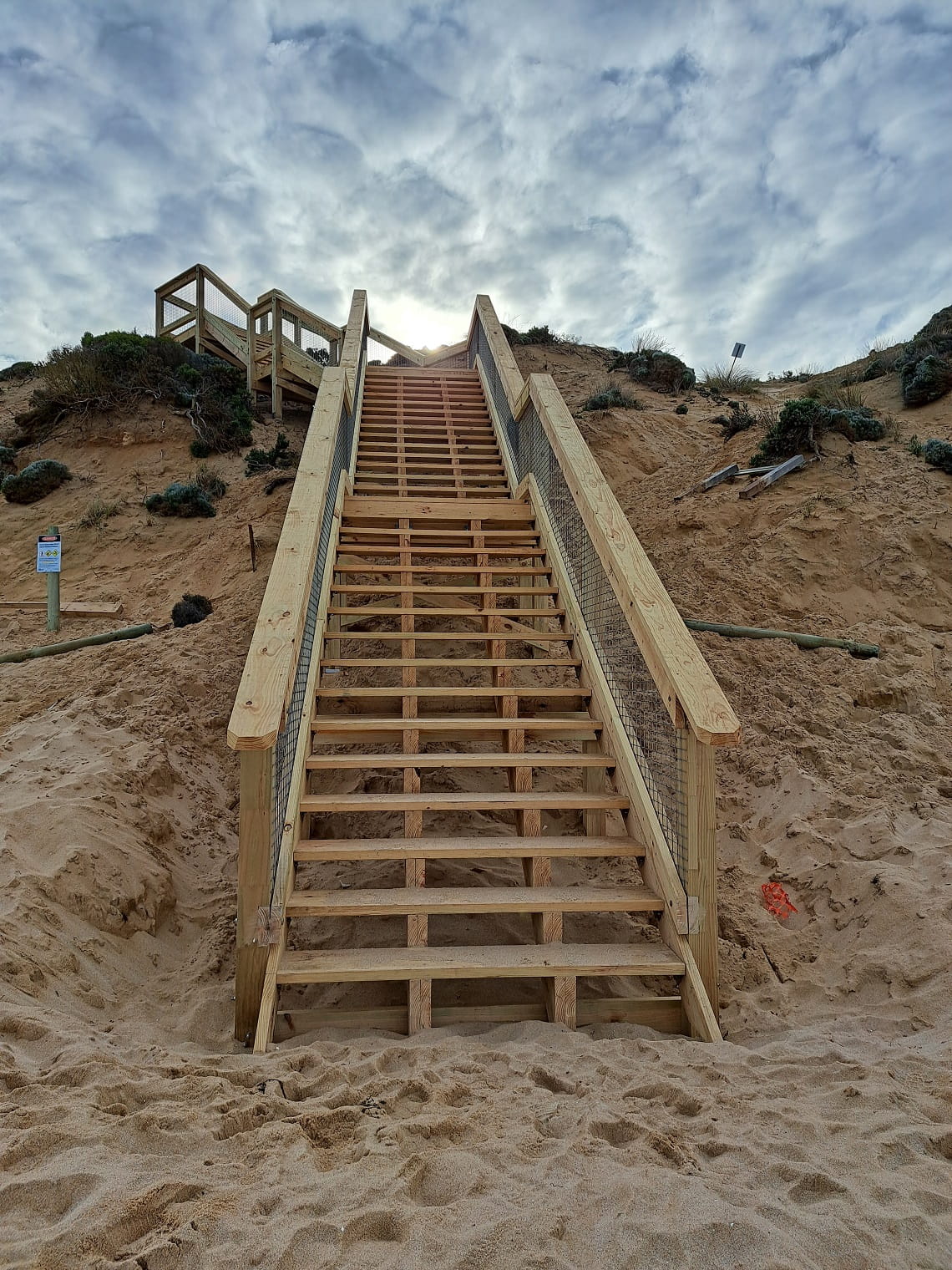 A portrait photo of the new stairs at Number 16 Beach. The photo is taken from the bottom of the stairs, looking up from the beach to the top of the stairs, which leads to grassy sand dunes.