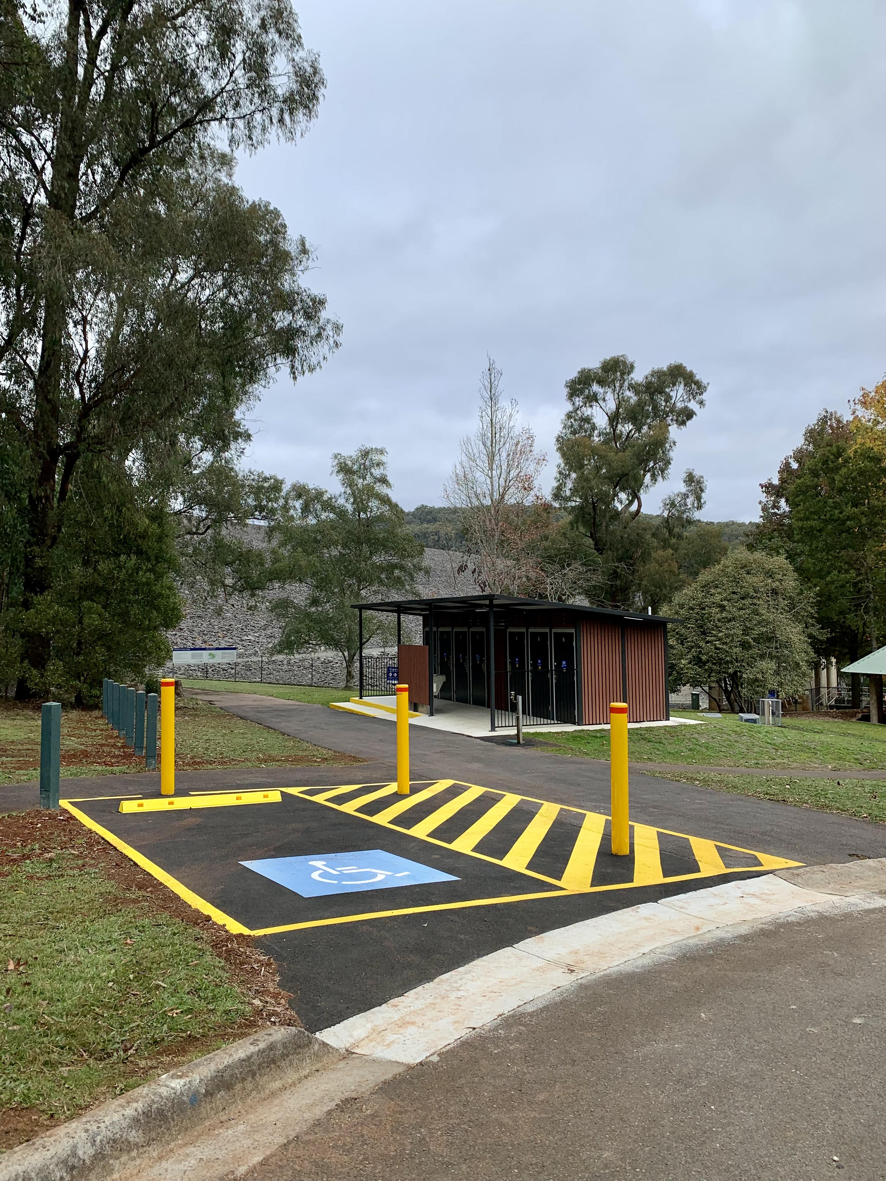 A DDA-compliant carpark with bright yellow linemarking, and a timber-clad toilet block in the background.