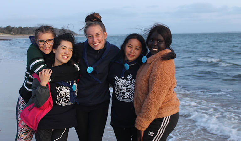 A group of five female friends hugging and posing on the beach.