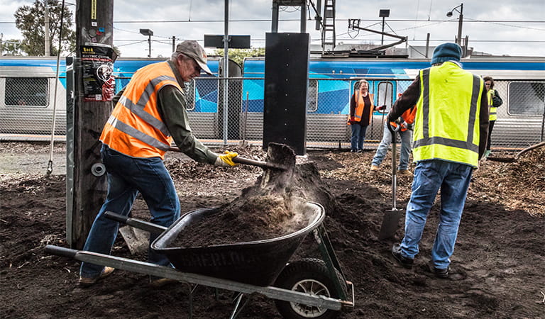 People in safety vests digging and transporting soil with a train station and train in the background.