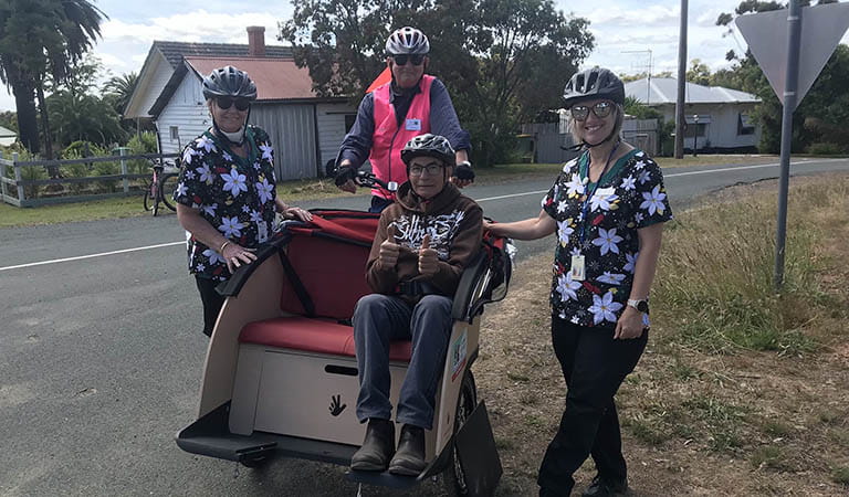 A person in a helmet and pink safety vest rides a TrioBike, with another person sitting on the padded seat of the TrioBike holding two thumbs up, while two smiling and helmeted women stand next to the TrioBike.