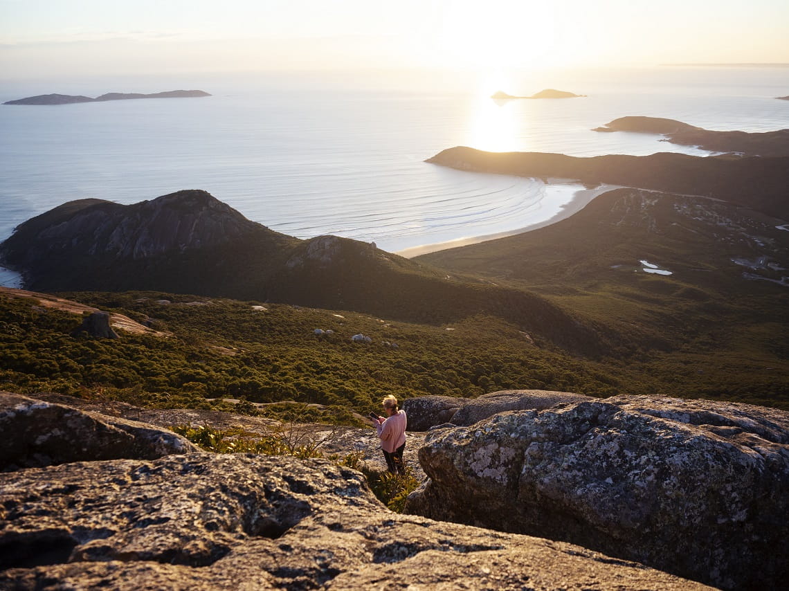 Woman standing on the top of a rock looking down to Wilsons Prom coastline as the sun sets