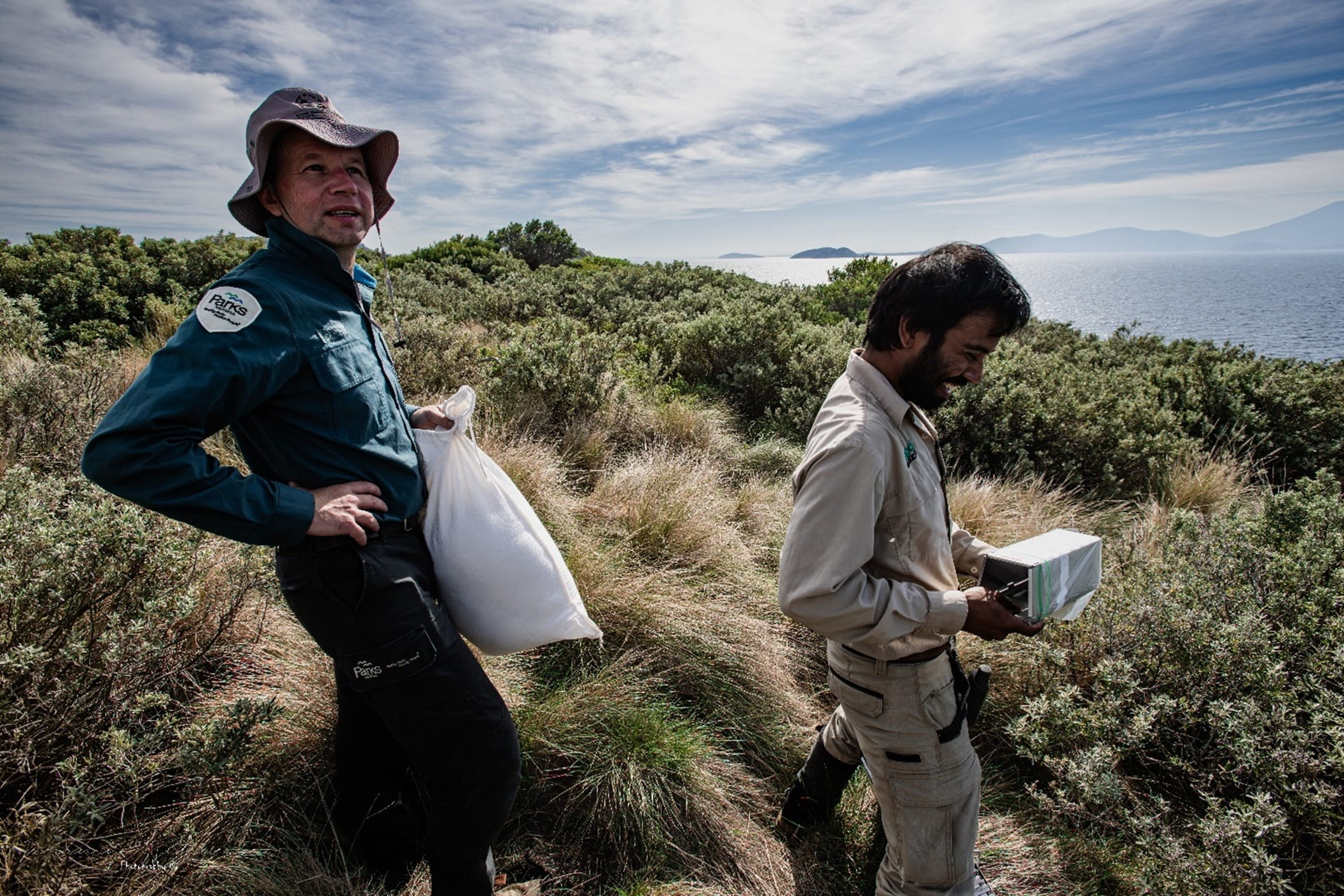 Two people stand with traps in their hands against a grass strewn backdrop.