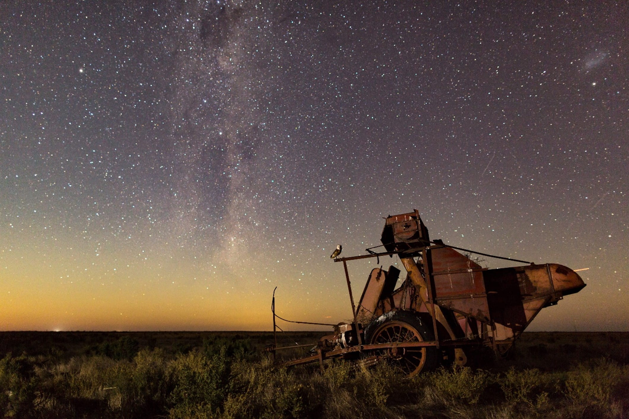 A starry sky, with a yellowing horizon, is illuminated behind a piece of farm equipment