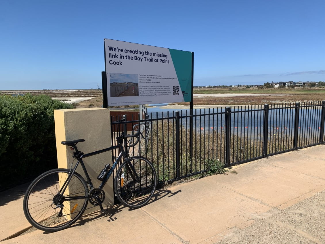 A bike leans against a railing with a Victorian Government sign saying "we're creating the missing link in the Bay Trail at Point Cook".
