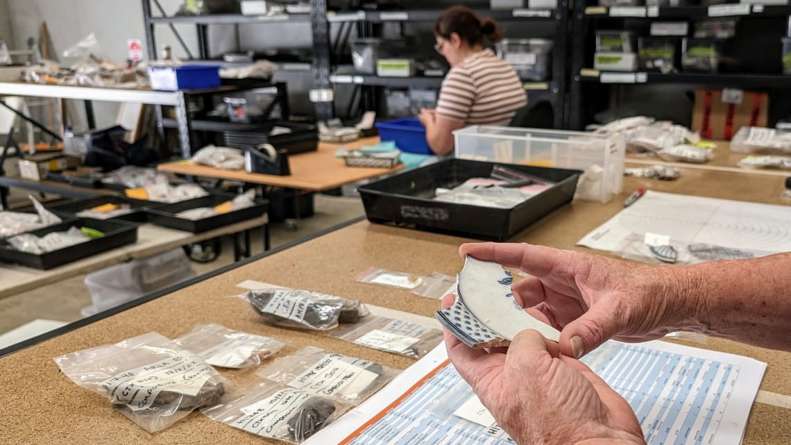 A table covered with bits of pottery and other items: an archaeologist hold a piece of a ceremic plate