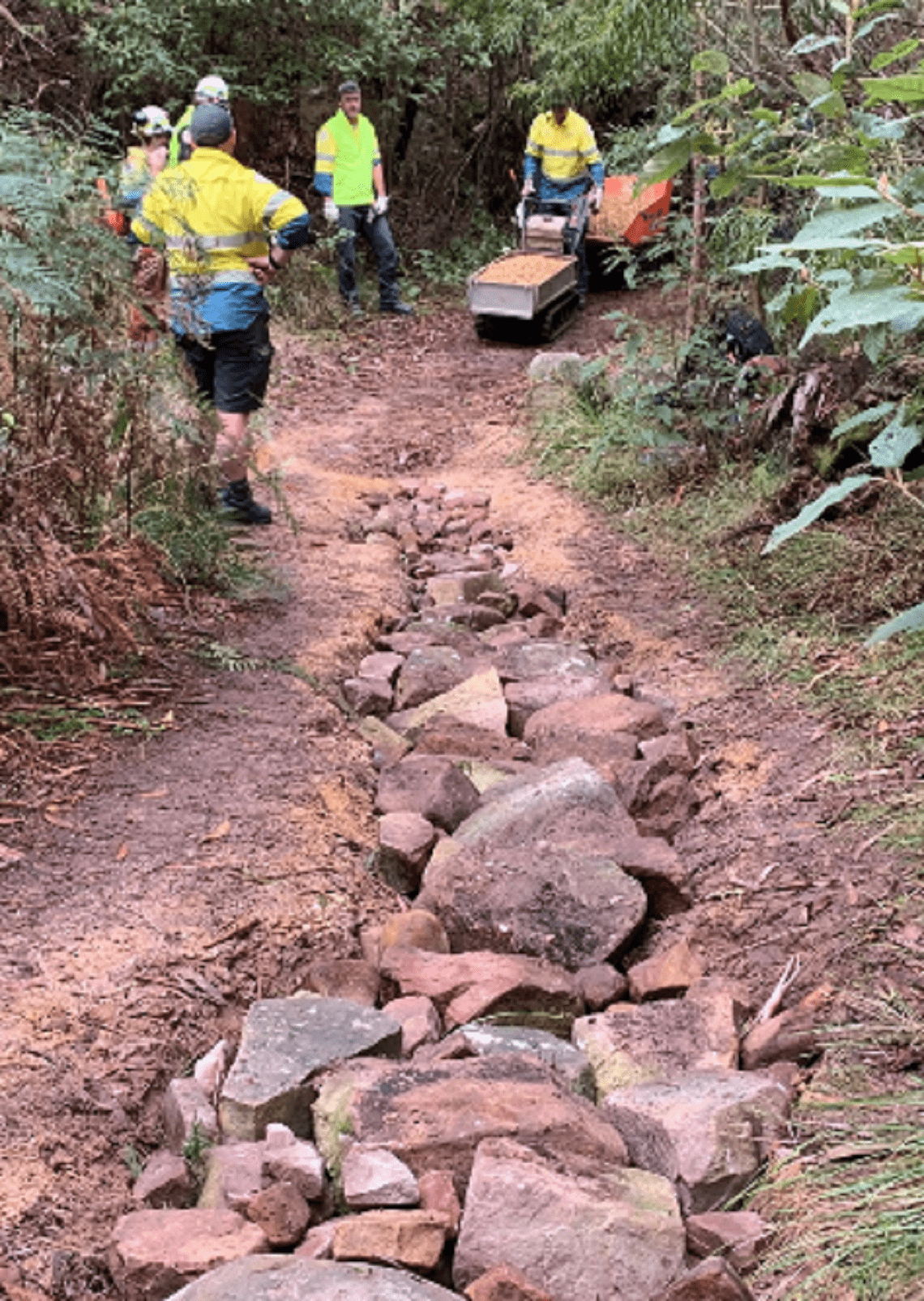 Image shows a team in high vis clothing standing beside a track with a deep erosion in the centre that has been filled with larger rocks before covering.