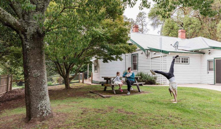 A mother sits at a table with her younger daughter and watches her older daughter doing a cartwheel on the lawn of the residence at Buchan Caves Park.