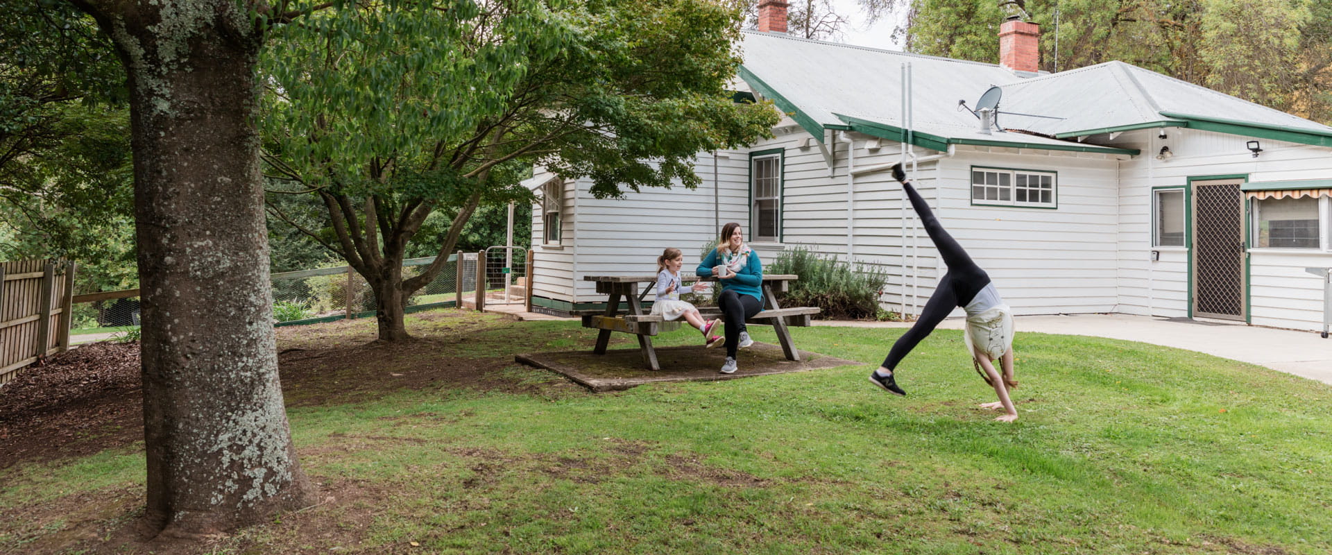 A mother and daughter seated at a picnic table cheer a young girl performing a handstand on the grass outside Caves House.