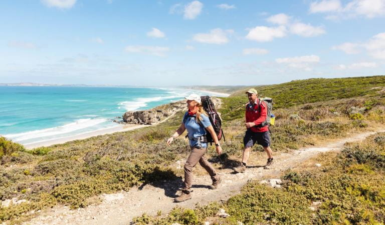 A man and woman hike on a rocky coastline covered with short green shrubbery and blue ocean wave-break in the distance. 