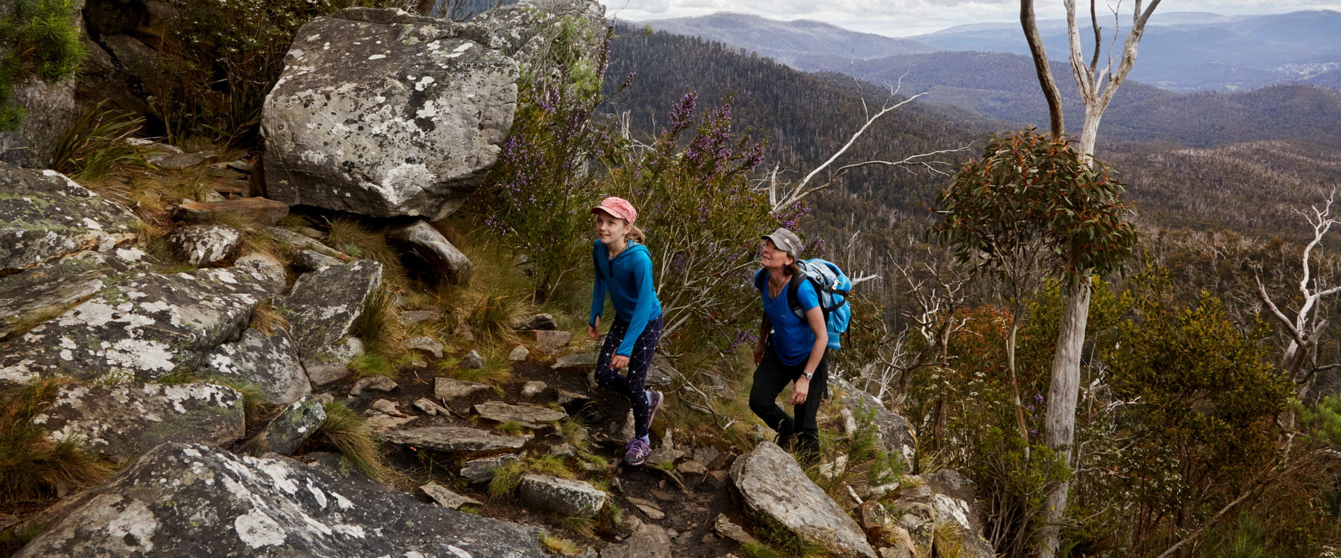 A woman and young girl hike up a rocky trail with a vast scenic mountain view stretching out into the distance behind them.