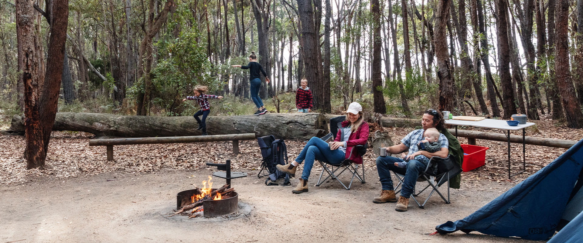 Mother, father and infant sit in camp chairs around a campfire while their other children play on a fallen log nearby
