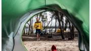 A man in a yellow jacket holding a cup of tea walks towards his partner in a tent 