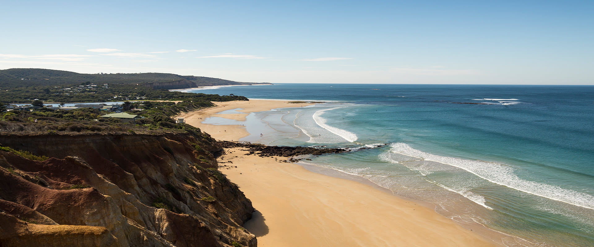 A view down the coastline from a lookout with the a sandy beach between the ocean of the right hand side and sandstone cliffs on the left.