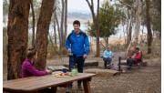 A man brings a kettle to the picnic table where his partner is sitting, while an older couple sit around a campfire in the background.