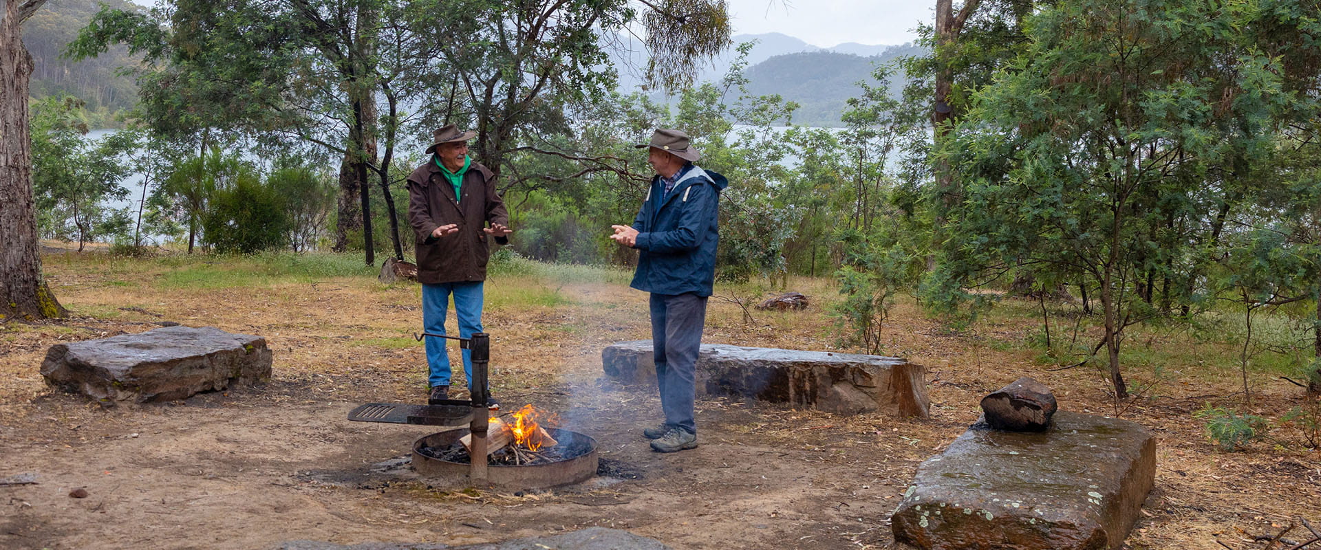 Two people stand over a campfire in a forested camping area. There are seats built into group. A large lake in the background.
