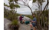 A man and women stop for a drinks break next to granite rocks on the Southern Circuit hiking trail at Wilsons Promontory National Park