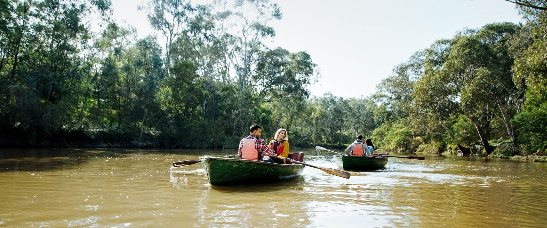 Two pairs of people row boat down a river with tall trees on its banks. 