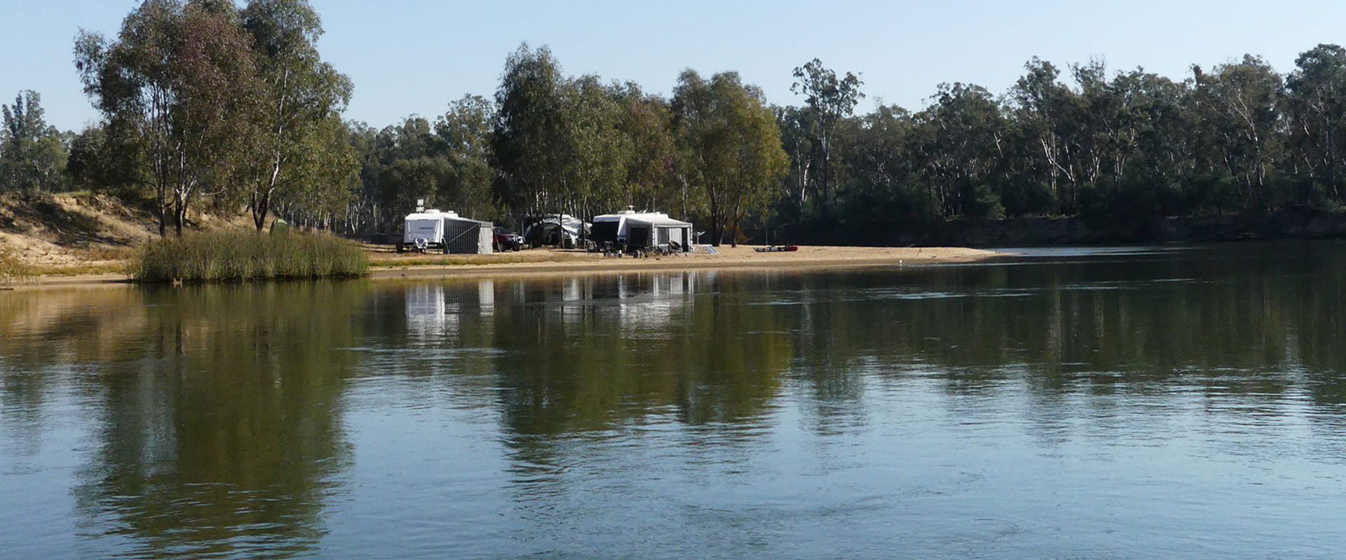 Caravans and Tents are setup on a sandy foreshore of a river and lake surrounded by rugged bushland.