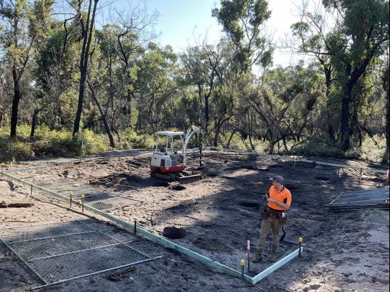 Early works shows white pegs and markers being laid out by a person in high vis on a sandy soil cleared construction site. There is a small digger working in the background stand tall native trees and blue-sky shines brightly above