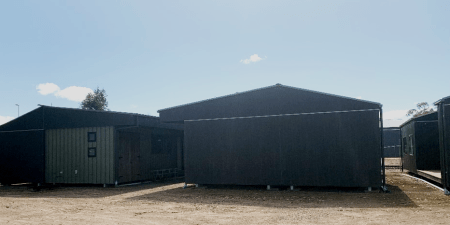 Back and front view of two cabins with a third cabin showing a built-in outdoor seating area. The cabins have black roofs and green coloured timber cladding. There are black framed windows in the walls.