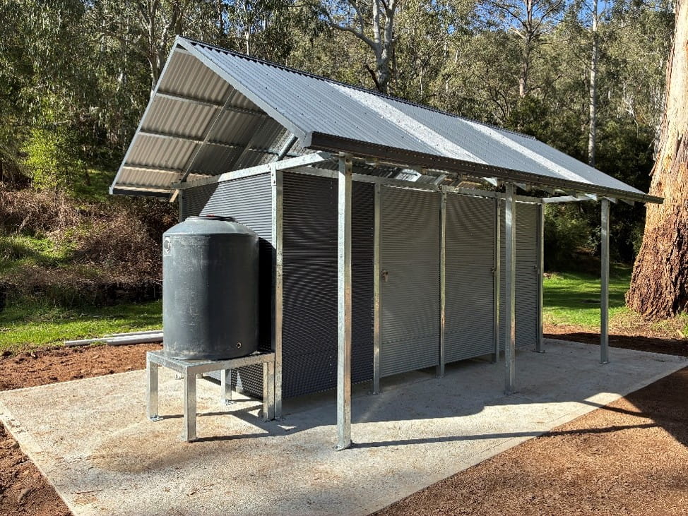 Modern accessible toilet block at Sheepyard Flat with corrugated steel walls and roof, concrete slab flooring, and a rainwater tank. The facility is surrounded by grassy open space and tall eucalyptus trees in a bushland setting.