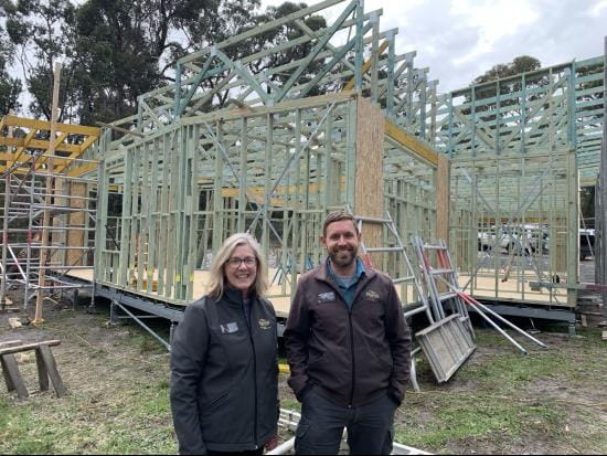 Two Parks Victoria team members are smiling standing in front of the timber frame of the new Cape Conran office building. There is green grass in the foreground, tall trees behind and the sky above is cloudy but bright