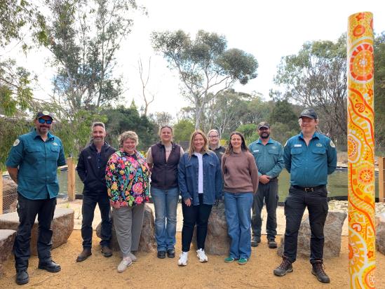 Group of nine people smiling standing in front of display pond. There is a pole at right wrapped with a First Nations artwork of bright colours of yellow, orange and white. Behind the groups stand tall native gum tress