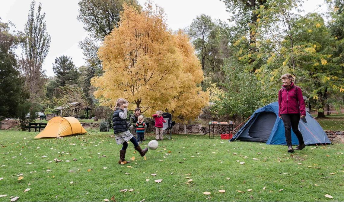 A mother and daughter kicks a football in front of dad and two younger children in front of tents at Buchan Caves Reserve.