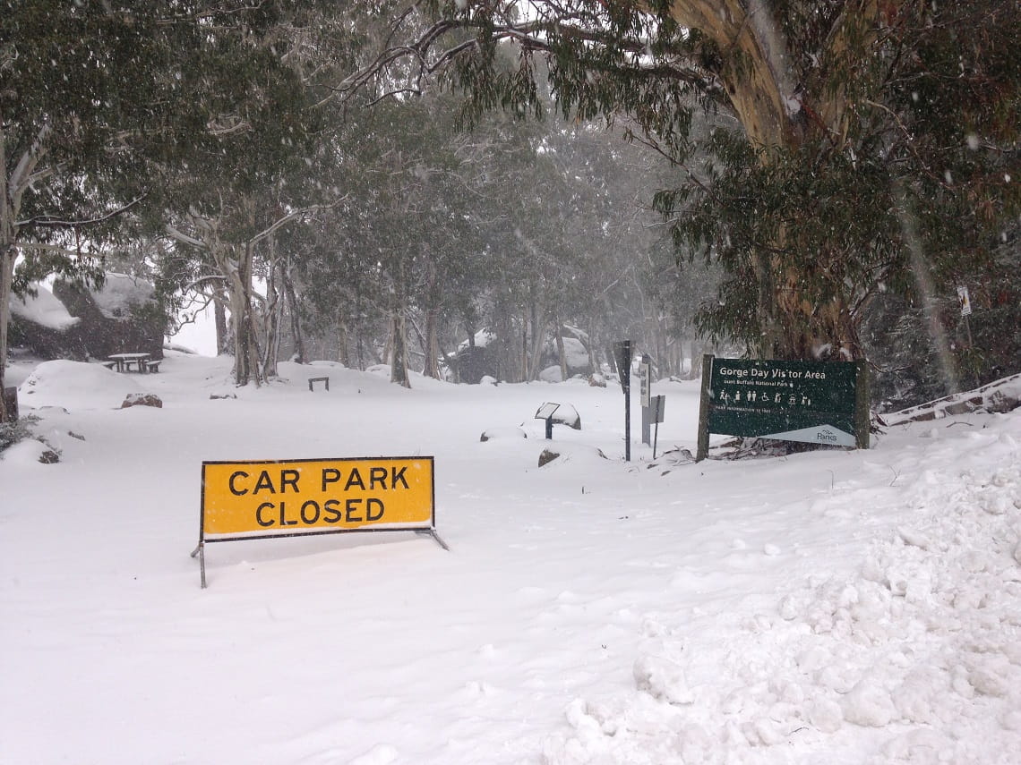 Snow-covered car park surrounded by tall trees. Yellow sign stating 'CAR PARK CLOSED'. Another sign indicating this is the Mount Buffalo Gorge Day Visitor Area.