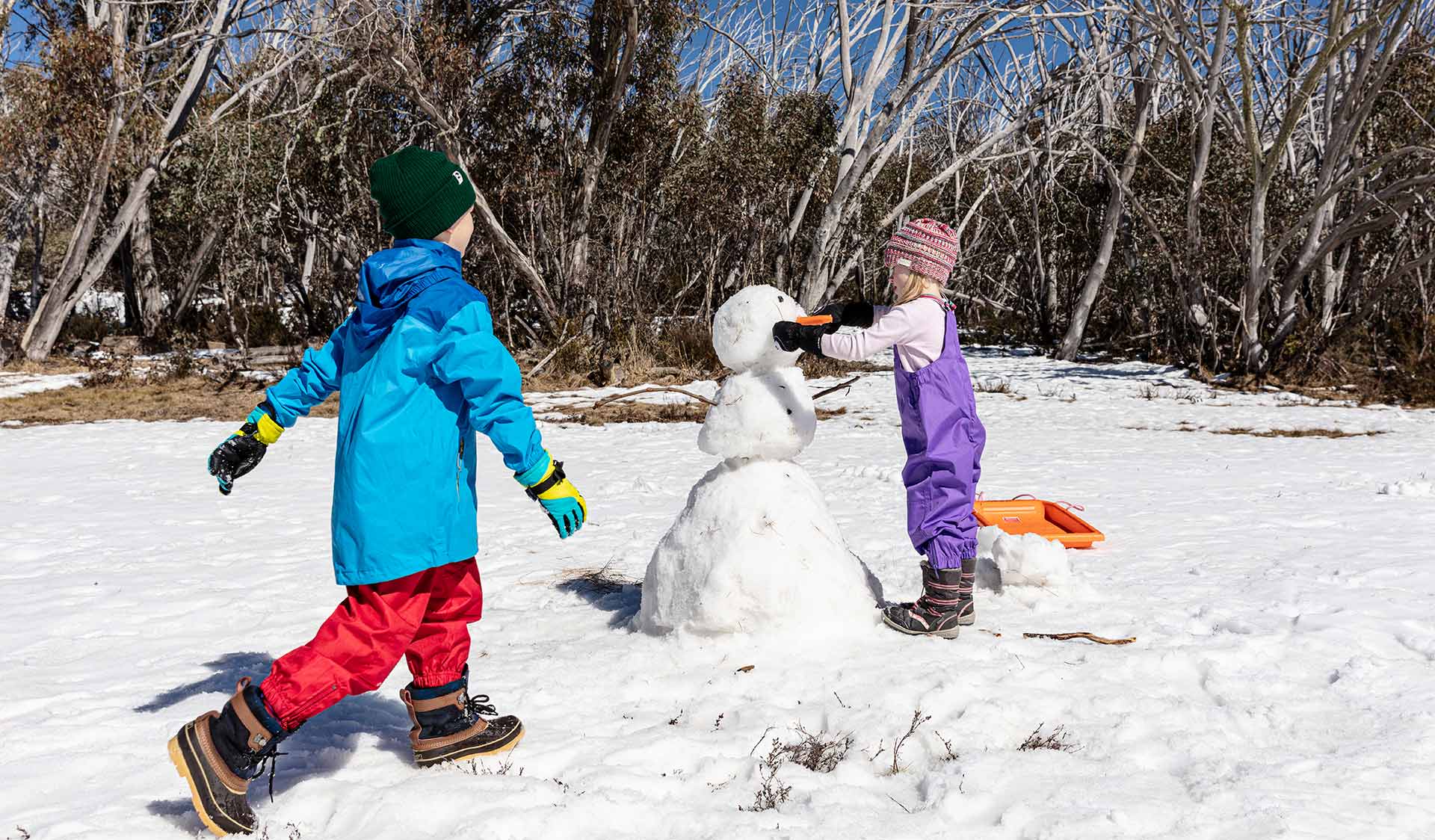 A young girl puts the final touches on her snowman as she is joined by her brother
