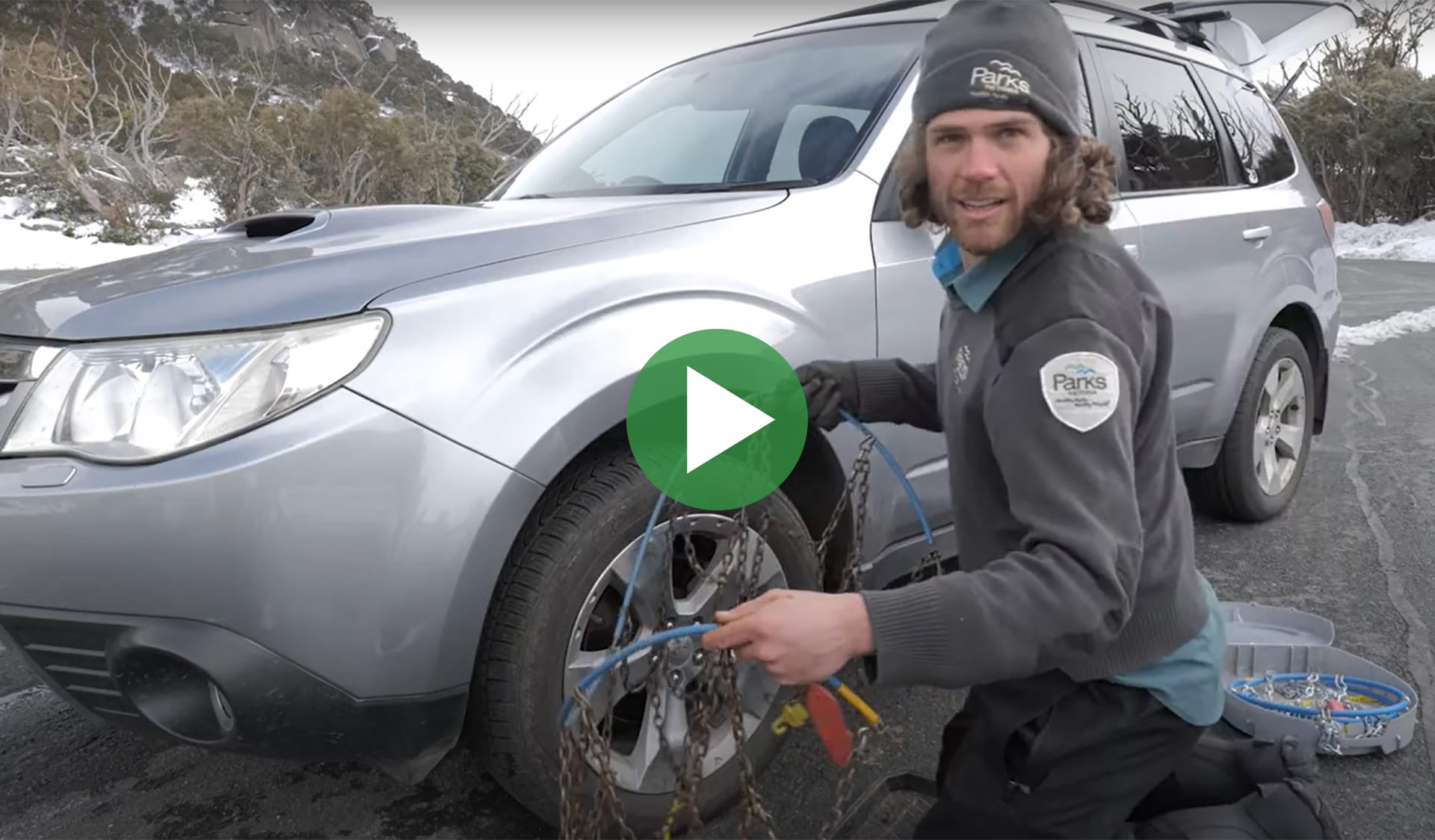 Video still of a Parks Victoria staff member in uniform demonstrating how to fit snow chains on a car with snow in the background.