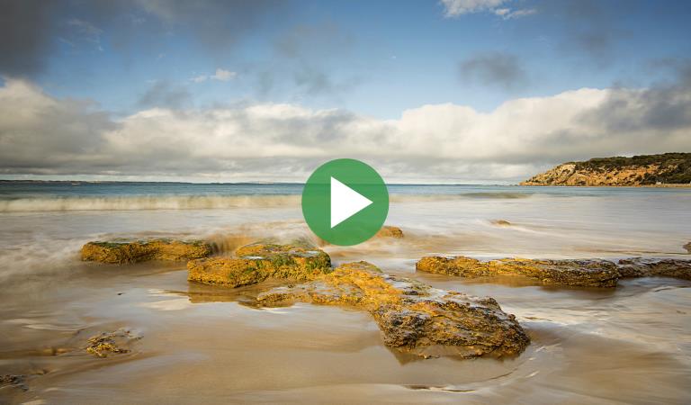 Waves crashing over rocks on the beach with video play icon overlay