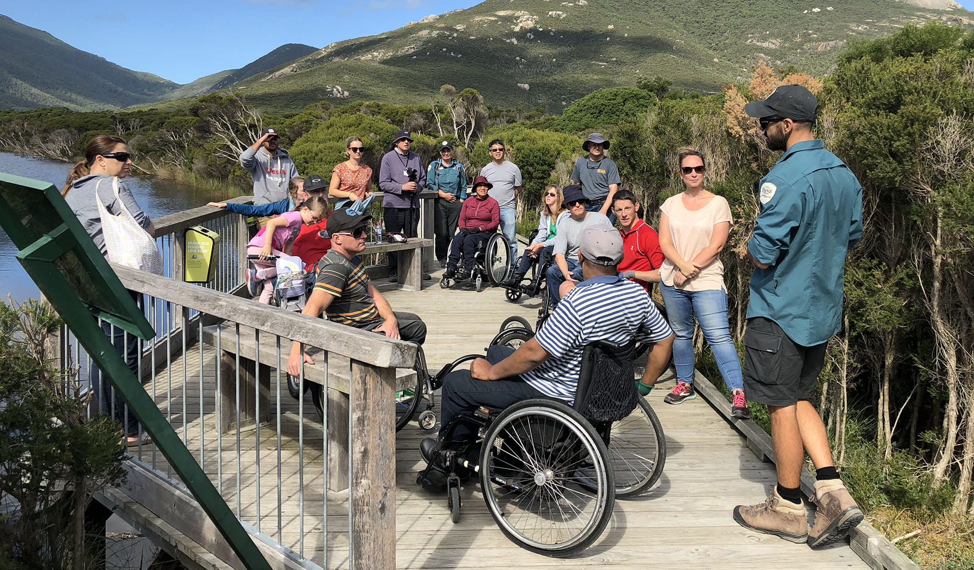 A group of people with various physical abilities gathered with rangers on a boardwalk beside a river with bushland and a mountain in the background. 