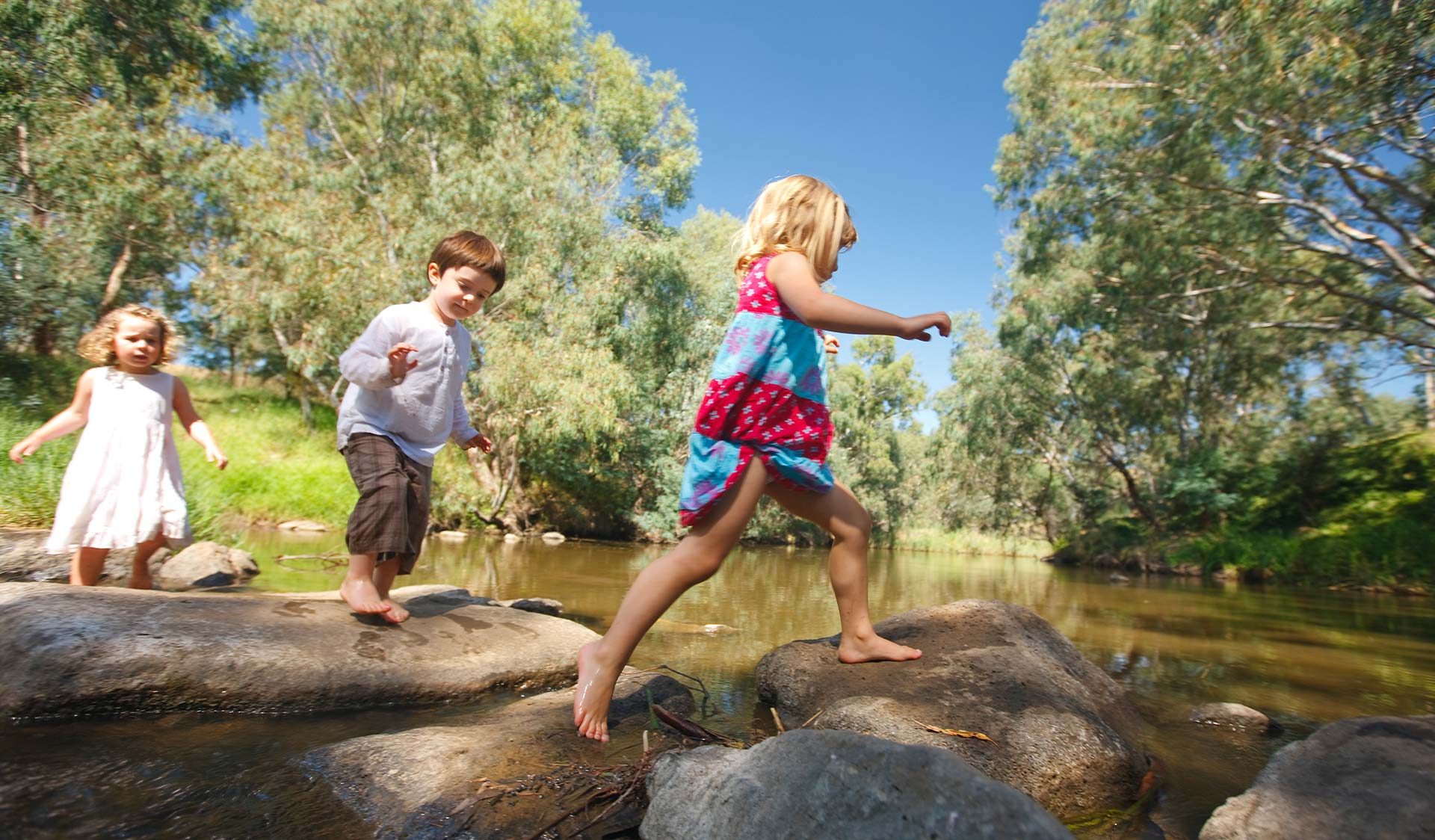 Three young children walking on rocks across a river