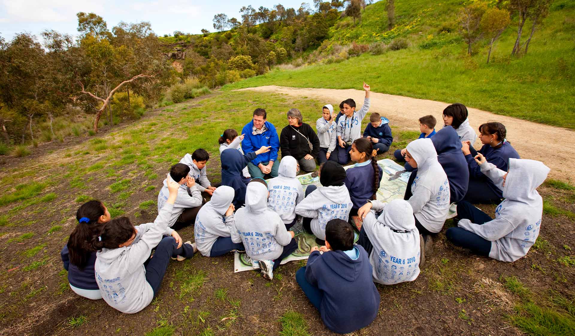 $name Group of school students in uniform sitting on grass engaging in discussion with teachers