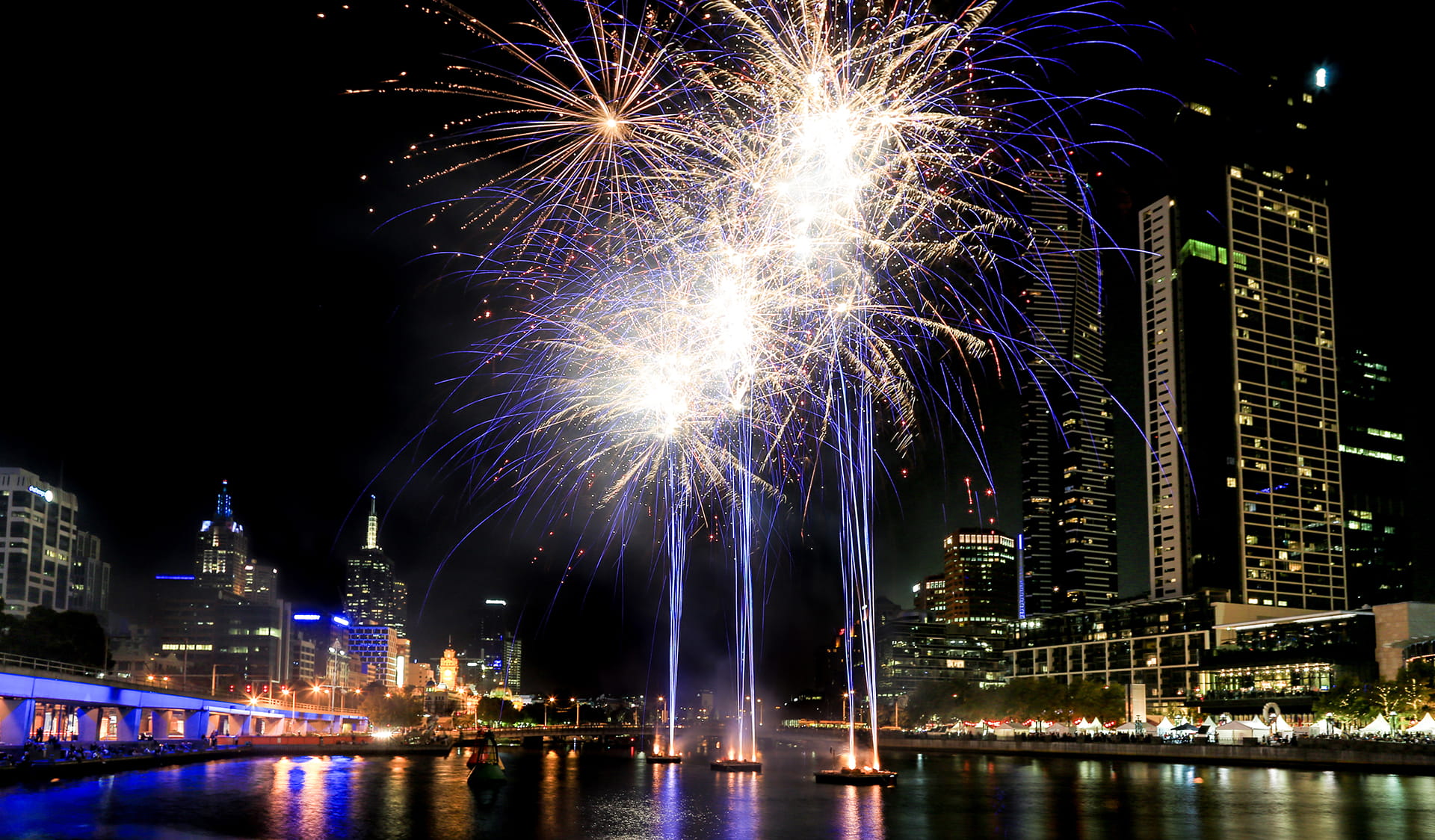 Fireworks display over a river running between city buildings.