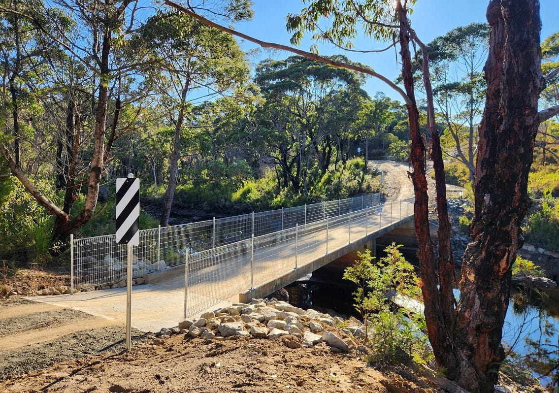 A concrete bridge with railing extends over a small river, surrounded by trees.