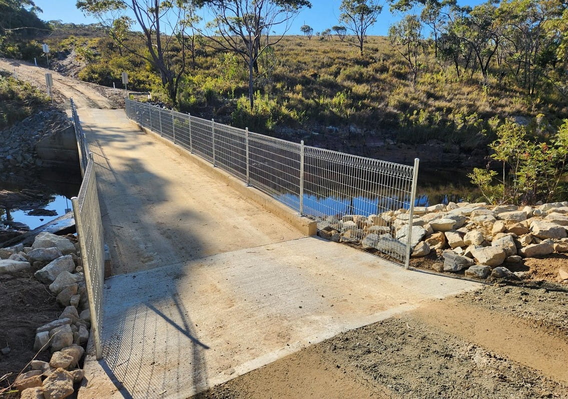 A concrete bridge with metal handrails crosses a river onto a gravel road, surrounded by bushland. 