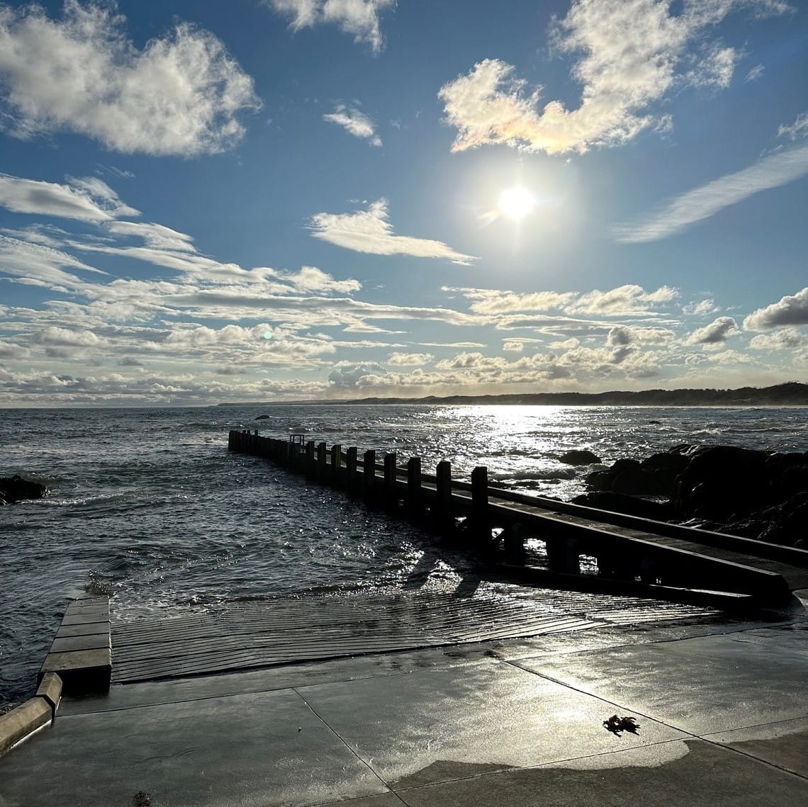 A concrete ramp declines into choppy waters, with a jetty in the distance and the sun setting over the shoreline.