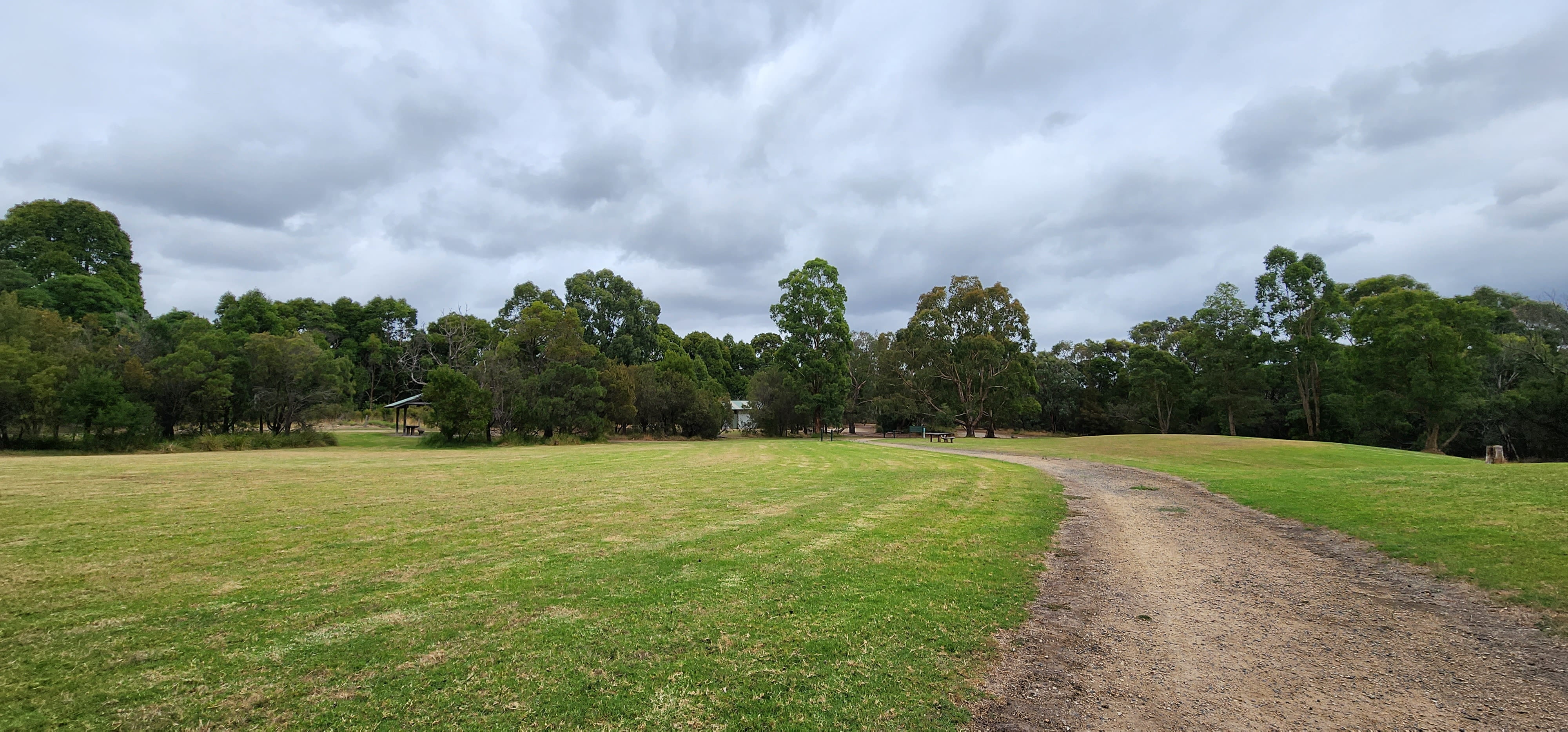 Image shows a gravel path winding in between two large grass fields. There are gums and green trees and a grey sky in the background.