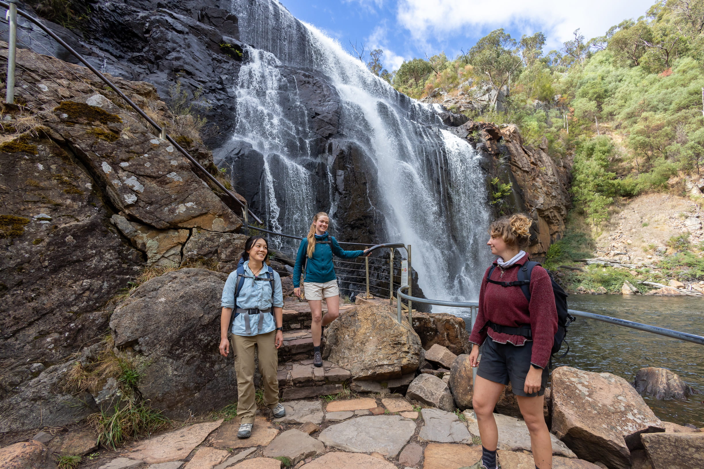 Three female walkers standing at viewing area on a track at the base of the waterfall, with the waterfall in the background
