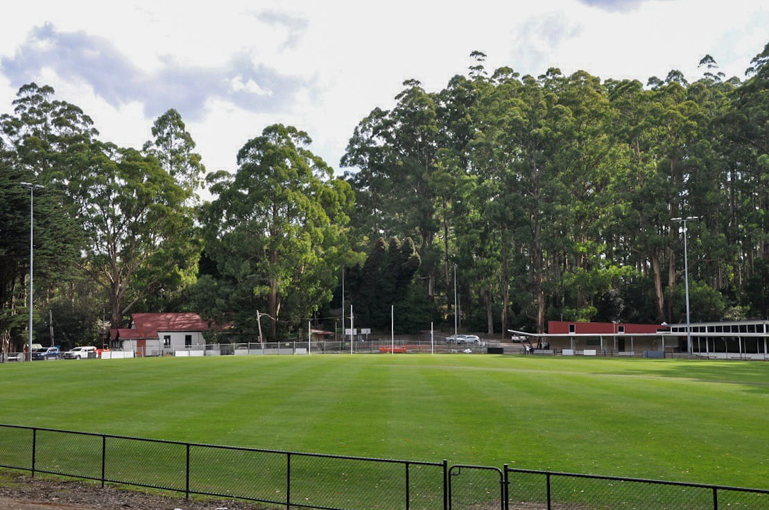 A wide landscape view of the Olinda Recreation Reserve. A green, grassy oval is in the foreground, with buildings, a construction site and tall trees in the distance. 