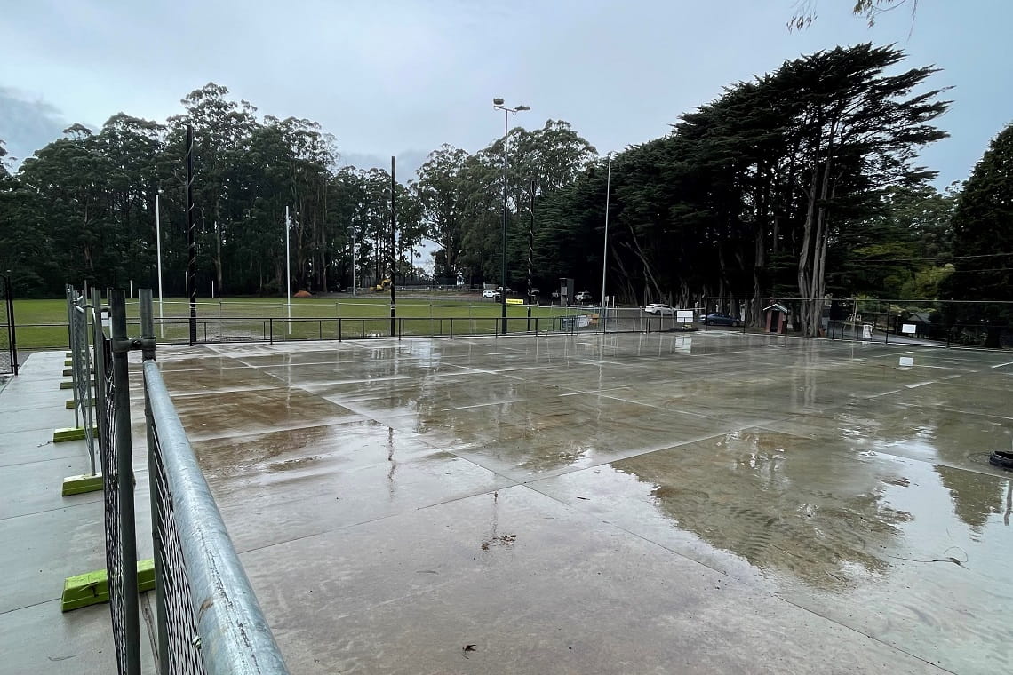 The construction of the new netball courts - a large concrete slab has been poured, and is currently surrounded by temporary fencing. In the background is the oval and large trees.