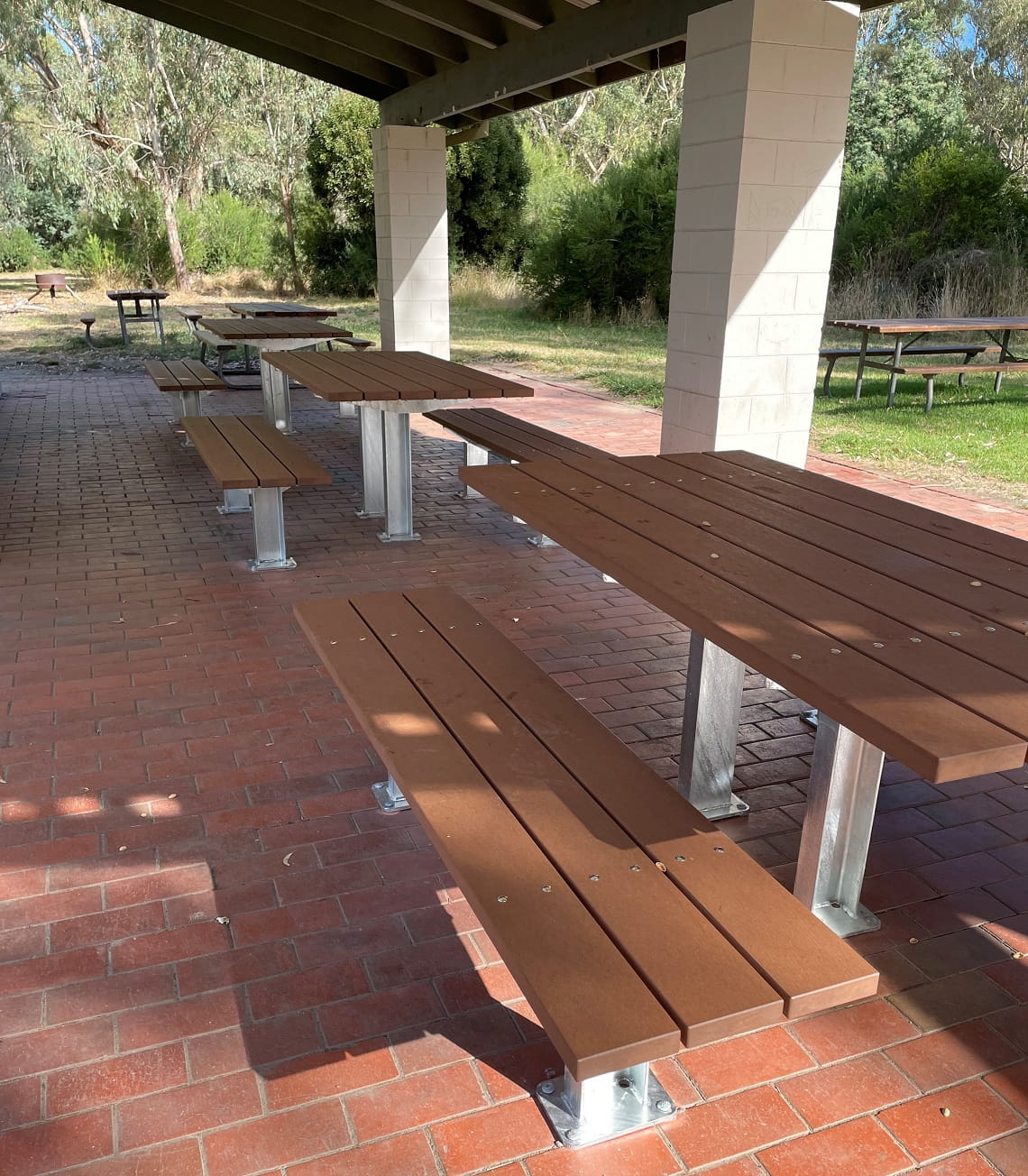 New picnic tables installed at Birrarung Park. There are three tables in a row, sitting on top of a red brick surface, underneath a large picnic shelter.